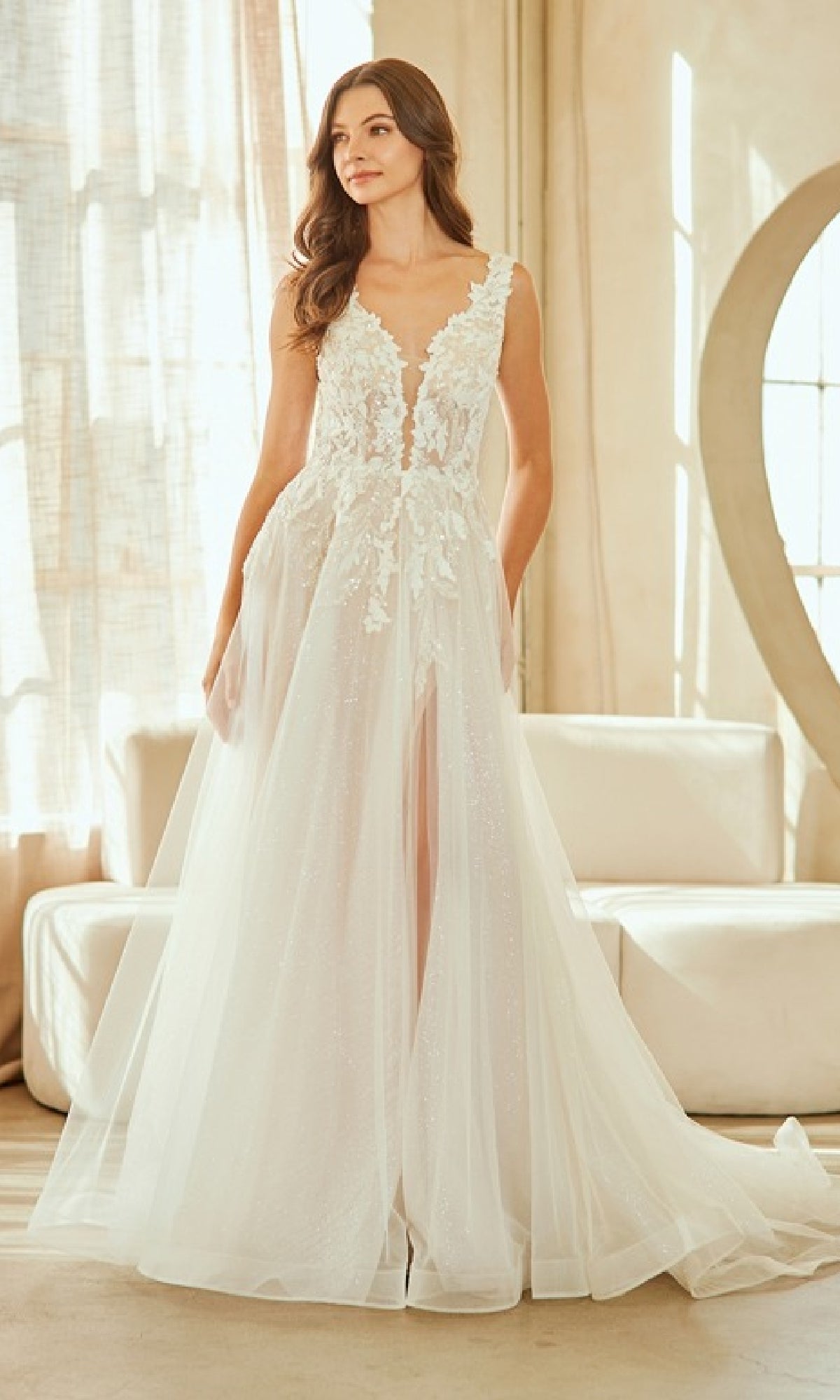 A woman stands indoors wearing the Amelia Couture 2140, a sleeveless, floor-length white tulle wedding dress with a deep V neckline, floral appliqué details, and an elegant corset bodice.